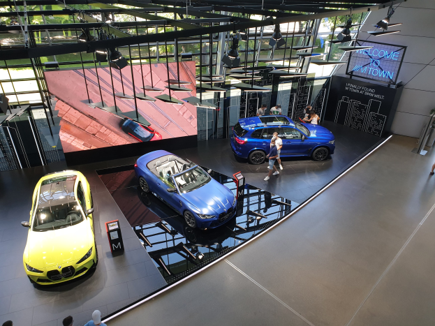 Eine Gruppe von Menschen, die im BMW Museum in München stehen, mit Autos auf der linken Seite und einem Schild mit der Aufschrift "BMW Museum München" auf der rechten Seite, mit Blick auf eine Außenansicht von Bäumen, Gebäuden und Himmel durch Glasfenster.