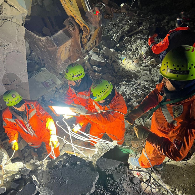 Eine Gruppe von Menschen in orangefarbigen Uniformen und grünen Helmen, die auf Schutt arbeiten, mit einem Kran im Hintergrund, der Rettungsgeräte und -materialien hält.