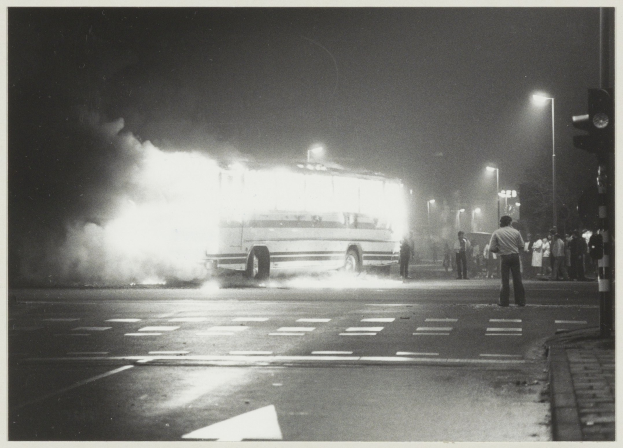 Ein Schwarz-Weiß-Foto eines brennenden Busses am Straßenrand mit einer Gruppe von Menschen auf dem Gehweg rechts und Hintergrundstraßenlaternen.