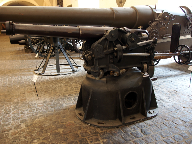 Eine große Gatling-Kanone mit langem Lauf und Seitengriff ist in einem Museum ausgestellt, umgeben von Bodenständen, mit einer fensterumrahmten Wand im Hintergrund, die an eine Burg erinnert.