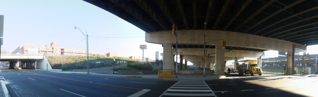 Ein Lastwagen fährt unter einer Brücke auf einer Stadtstraße, umgeben von Gebäuden, Straßenlaternen, Verkehrszeichen, Strommasten, Stromkabeln, Kraftfahrzeugen, Barrieren, Büschen, Bäumen und einem klaren blauen Himmel.