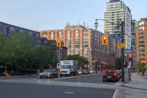 Eine belebte Stadtstraße mit mehreren Fahrzeugen, darunter ein Lkw, ein Auto und ein SUV, umgeben von Laternenmasten, Ampeln, Texttafeln, Bäumen und Gebäuden unter einem Himmel.