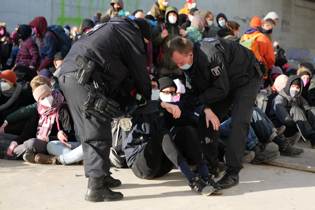 Eine Gruppe von Menschen, einige mit Mützen und Masken, sitzt auf dem Boden mit zwei Polizeibeamten in der Nähe, vor einer Wand mit Schrift, die auf eine städtische Umgebung hinweist.