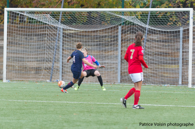 Eine Gruppe von Frauen, die Fußball auf einem grünen Feld umgeben von Bäumen spielt, mit einem Tor im Hintergrund und Text am unteren Rand des Bildes.