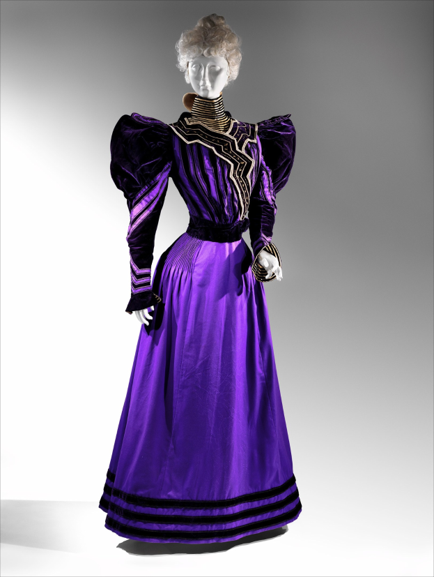 Eine Schaufensterpuppe in einem lila Abendkleid mit schwarzen und goldenen Mustern, mit einer eng anliegenden Taille, einem langen, bodenlangen Rock mit goldener Stickerei und Perlen und Spitze an der Taille, gestylt mit einem eleganten Dutt und zarten Ohrringen.