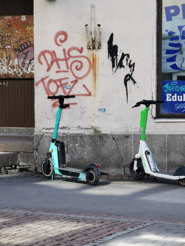 Zwei elektrische Roller nebeneinander auf einer Straße mit einer Graffiti-Wand und einem Plakat im Hintergrund geparkt.