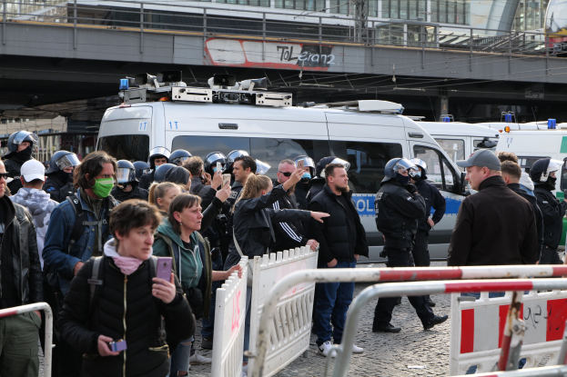 Eine Gruppe von Menschen steht vor Polizeifahrzeugen, einige tragen Helme und halten Telefone, mit Barrikaden im Vordergrund und einer Brücke und Gebäuden im Hintergrund in Berlin, wo die Polizei Demonstranten auseinandertreibt.