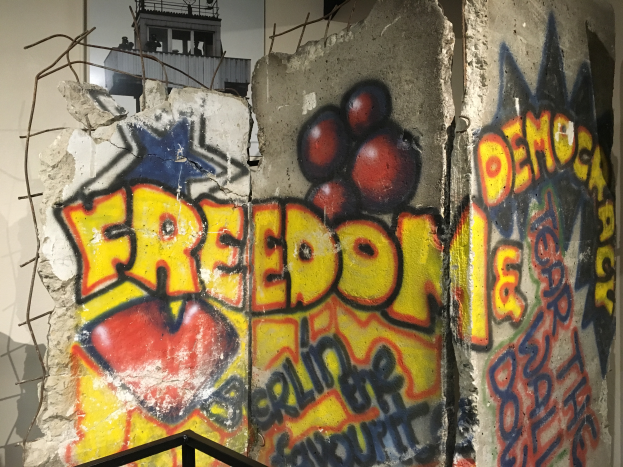 Eine Wand mit mutiger, schwarzer Graffiti, die 'Freiheit und Demokratie' lautet, in einem Raum mit einem Geländer unten und einem Gebäude mit Fenstern im Hintergrund.