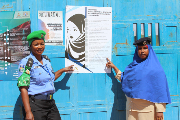 Zwei Frauen stehen nebeneinander vor einer blauen Tür, die mit Plakaten bedeckt ist, wobei eine eine blaue Jacke, schwarze Hosen und eine grüne Mütze trägt und die andere ein Blatt Papier hält, was darauf hindeutet, dass sie Teil einer Polizeikraft sind.