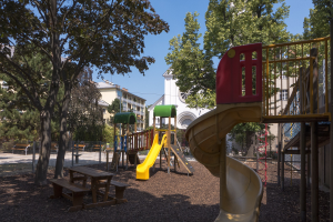 Spielplatz mit einer gelben und roten Rutsche, umgeben von Bäumen, Bänken, einem Zaun, Gebäuden und einem klaren blauen Himmel.