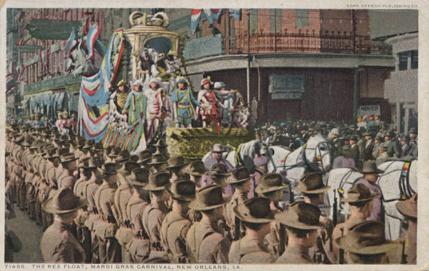 Eine alte Postkarte, die einen Mardi Gras-Umzug in New Orleans zeigt, mit Menschen auf Pferden, einem dekorierten Wagen mit Flaggen und Gebäuden im Hintergrund. Der Text unten lautet "The Rex Float, Mardi Gras Carnival, New Orleans".