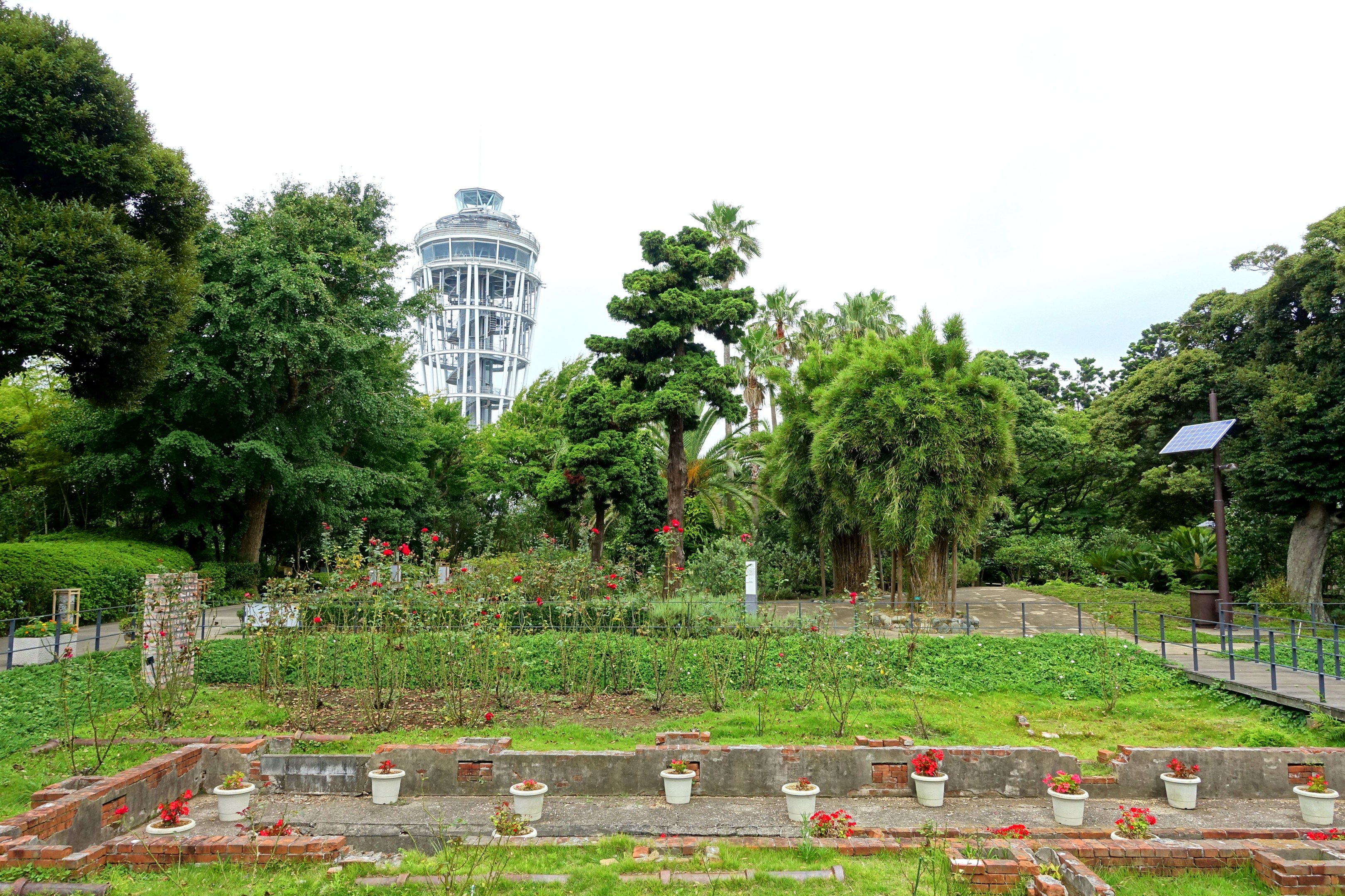 Ein Park mit einem Turm im Hintergrund, umgeben von grünem Gras, Pflanzen, Böumen, verstreuten Blumentöpfen, einer Straße mit Gelúndern und einem Pfahl mit einer Tafel, unter einem sichtbaren Himmel.