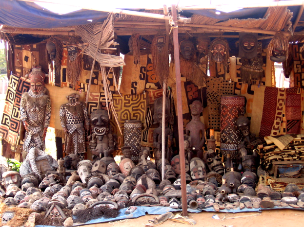 Ein Marktstand mit verschiedenen afrikanischen Masken, hölzernen Gegenständen, Kleidung und anderen Artikeln im Vordergrund, mit Bäumen und einem klaren blauen Himmel im Hintergrund.
