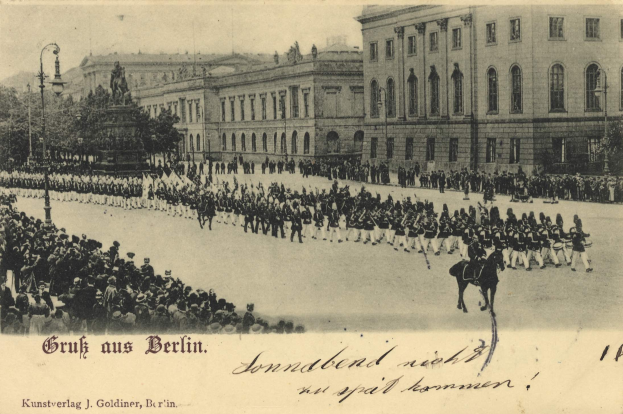 Ein altes Schwarz-Weiß-Foto einer Parade in Berlin, Deutschland, mit Menschen auf dem Boden und auf Pferden, einer Statue auf einem Podest, Straßenlaternen, Bäumen, Gebäuden mit Fenstern und einem bewölkten Himmel, mit Text am unteren Rand des Bildes.