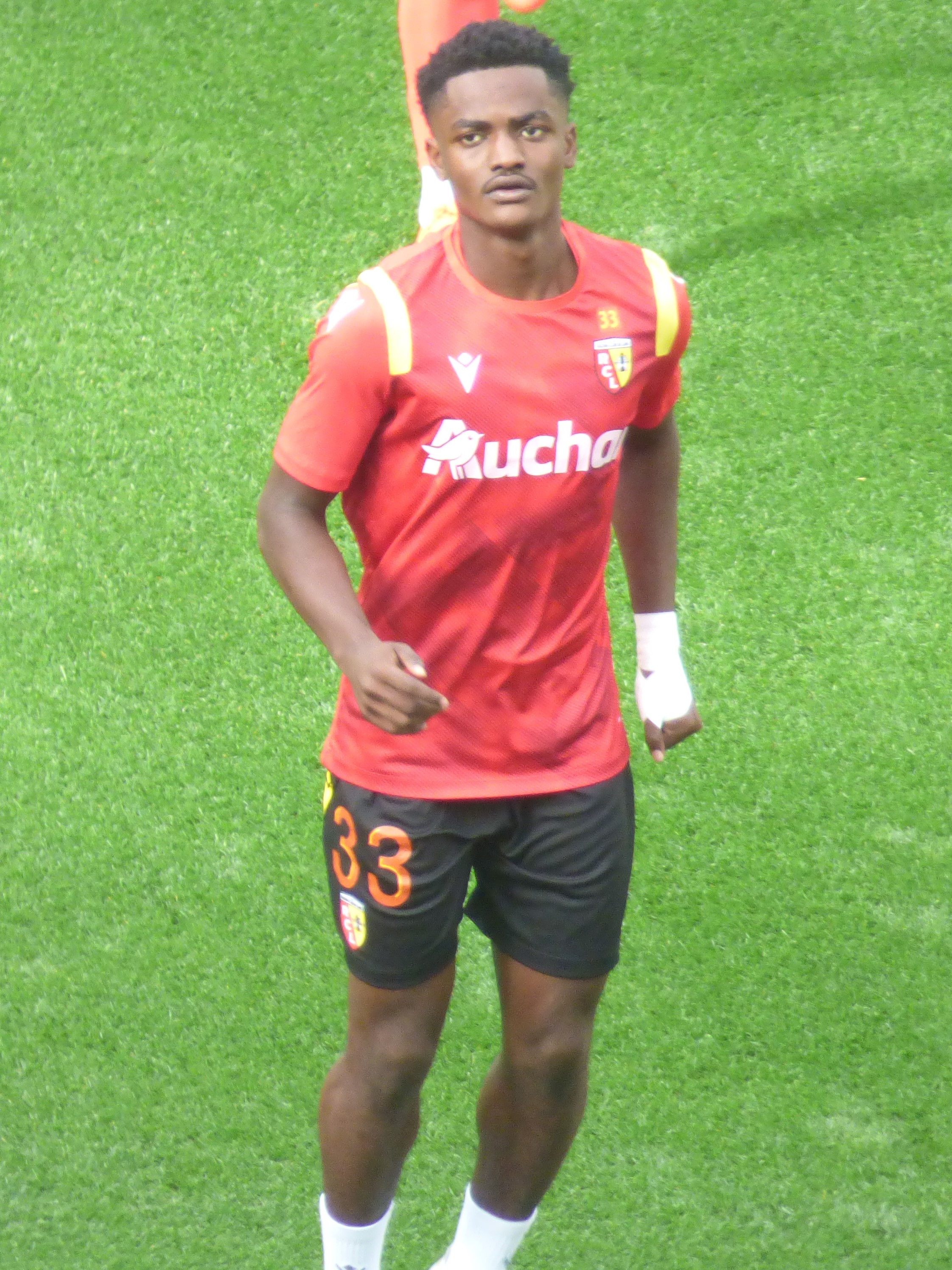 Emmanuel Adebayor, ein Mann in einem roten T-Shirt und schwarzen Shorts, geht auf einem Fußballfeld mit zwei Personen im Hintergrund.