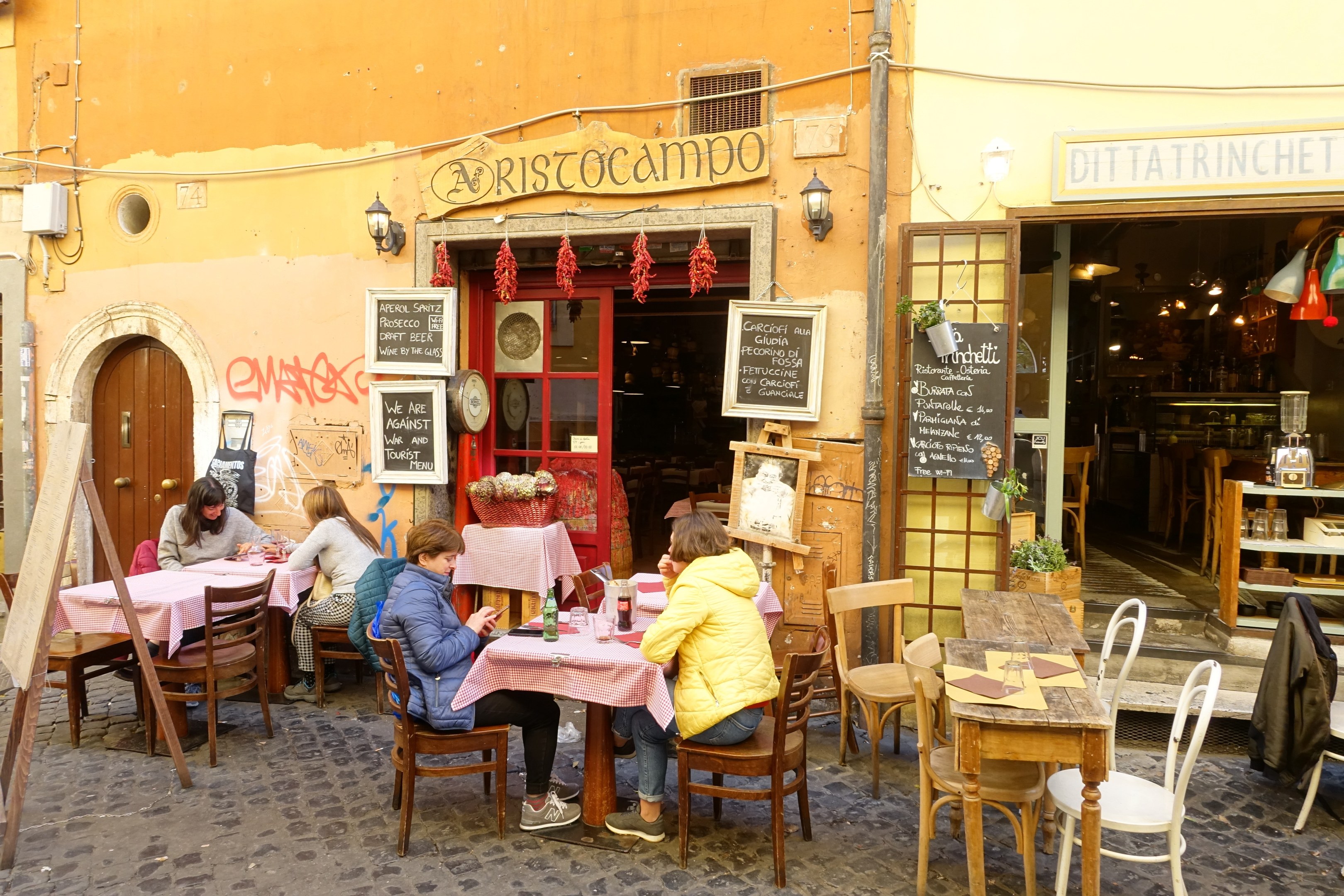 Eine Gruppe von Menschen an Tischen eines Restaurants in Rom, Italien, mit Flaschen, Gläsern und Dekoration, sowie beleuchteten Wänden mit Texttafeln.