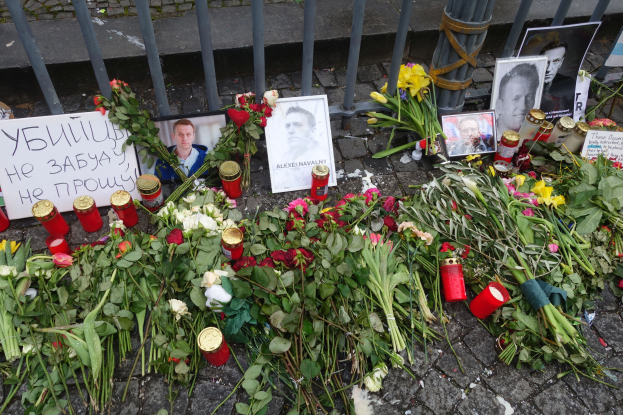 Ein Denkmal in Kiew, Ukraine, mit Blumensträußen, Kerzen, Fotoeinrahmungen und anderen Gegenständen, die auf dem Boden vor einem Zaun abgelegt sind, mit einer Straße im Hintergrund.