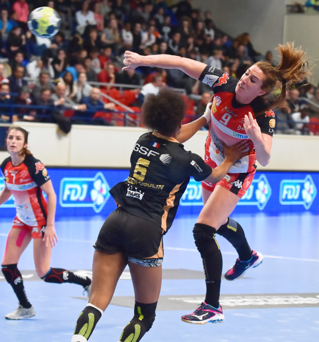 Frauen beim Handballspielen auf einem Feld mit einem Ball in der Luft, in einer Arena mit Zuschauern im Hintergrund.