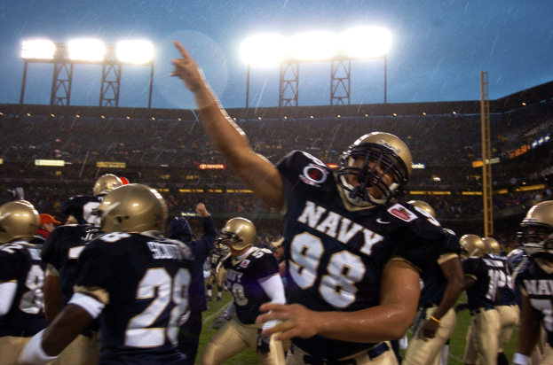 Navy-Fußballspieler feiern ein Touchdown auf einem regennassen Feld, tragen Helme und Uniformen, mit Zuschauern und Stadionbeleuchtung im Hintergrund.