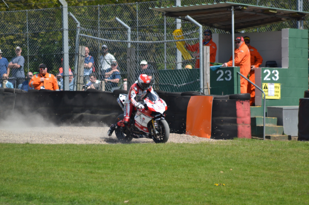 Eine Person in Helm und Handschuhen fährt ein Motorrad auf einem grasbewachsenen Feld, mit Zuschauern, Absperrungen, Treppen und einem nummerierten Schuppen im Hintergrund.