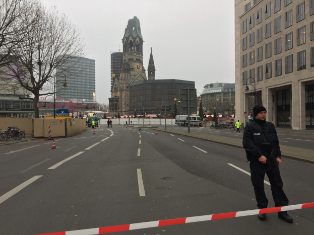Ein Polizist in schwarzer Uniform und Mütze steht auf einer Straße in Berlin umgeben von Verkehrskegeln, Fahrrädern, Laternenmasten, Bäumen und Gebäuden, mit einem Uhrenturm und einem klaren blauen Himmel im Hintergrund.