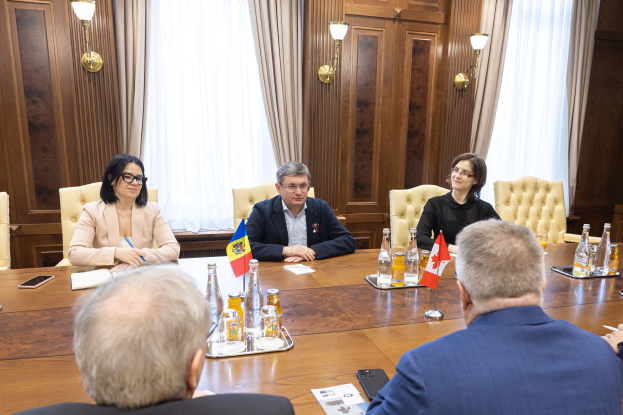 Eine Gruppe von Menschen sitzt um einen Holztisch in einem formellen Setting und diskutiert die Europäische Union mit verschiedenen Gegenständen auf dem Tisch, darunter Flaggen, Flaschen, Gläser und Papiere.