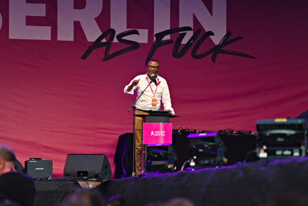 Mann in weißem Hemd und braunen Hosen auf einer Bühne mit einem pinken Banner 'Berlin as Fuck' und einer Menge davor.