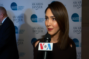 Emmy Rossum in einem schwarzen Kleid mit einem Mikrofon, mit einem Mann zu ihrer Linken und einem Banner mit der Aufschrift 'Aam Awards Dinner' im Hintergrund.