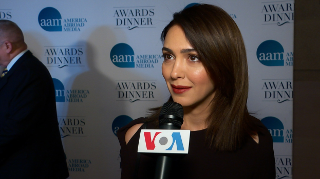 Emmy Rossum in einem schwarzen Kleid mit einem Mikrofon, mit einem Mann zu ihrer Linken und einem Banner mit der Aufschrift 'Aam Awards Dinner' im Hintergrund.