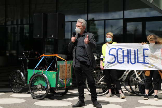 Ein Mann mit einer Maske hält ein Mikrofon vor einer Gruppe von Menschen, einige halten eine Fahne mit Text, Fahrräder, ein Fahrzeug mit einer Kiste und Lautsprecher, in einer öffentlichen Fläche mit einem Glaswand-Gebäude im Hintergrund.