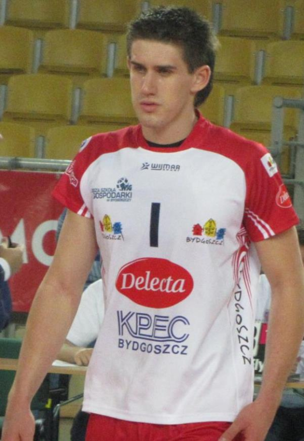 Ein Mann in rot-weißer Handballuniform steht auf einem Feld, entschlossen, mit Zuschauern und einem roten Banner mit der Aufschrift "Krasnoyarsk vs Bydgoszcz" im Hintergrund.