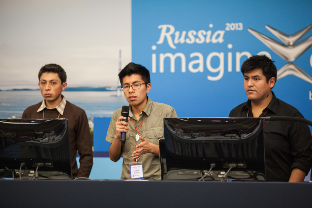 Drei Jungen stehen neben einem Schreibtisch mit zwei Computern, einer spricht in ein Mikrofon und trägt ID-Karten mit roten Markierungen, vor einer Wand mit einem 'Russia Imagine 2013'-Plakat.
