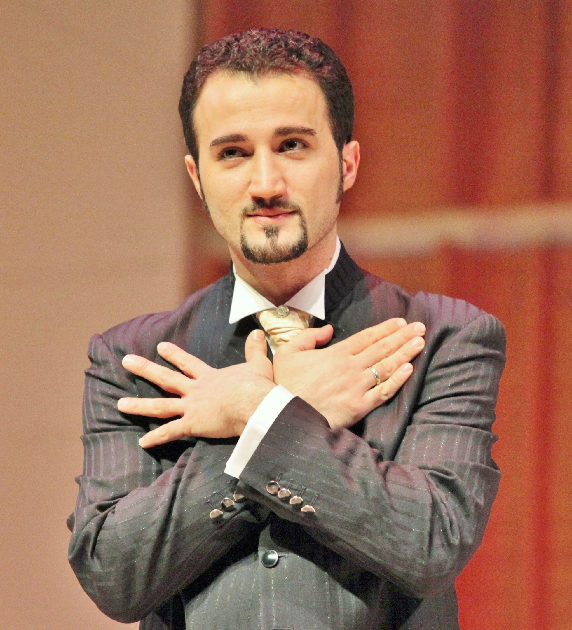 Ein Mann in Anzug und Krawatte steht selbstbewusst auf einer Bühne mit einer Wand im Hintergrund und sieht entschlossen aus.