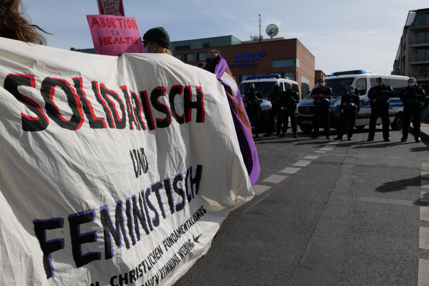 Gruppe von Menschen geht die Straße entlang und hält eine 'Solidarität und Feminismus'-Plakette, mit geparkten Fahrzeugen und Gebäuden im Hintergrund unter einem klaren blauen Himmel.