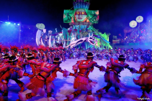 Eine Gruppe von Menschen in farbenfrohen Kostümen tanzt auf einer Bühne vor einem sitzenden Publikum, mit Lichtern, Polen, Ballons und einem Bildschirm, der eine Person in der Hintergrund zeigt, unter einem sichtbaren Himmel, bei der Rio-Karneval 2014.