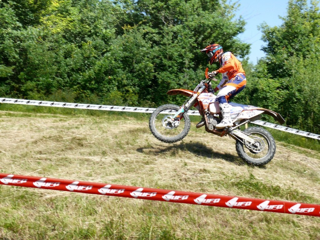 Eine Person mit Helm fährt ein Fahrrad innerhalb eines Absperrbandes mit Bäumen, Gras und Himmel im Hintergrund.