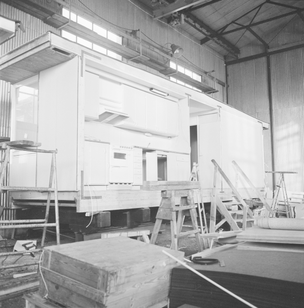 Ein Schwarz-Weiß-Foto eines Gebäudes im Bau mit Leitern, Holzboxen und anderen Gegenständen, die auf dem Boden verstreut sind, mit einer Wand mit Fenstern und Lichtern im Hintergrund.