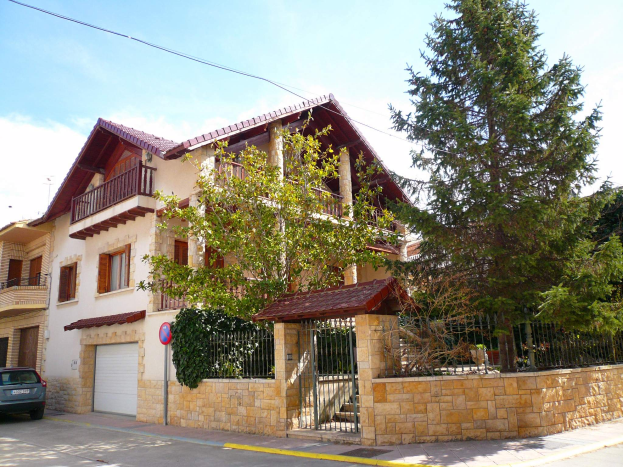 Eine Villa mit einem davor geparkten Auto, umgeben von einem Tor, einem Schild, einem Straßenschild, Bäumen und einer Wand, unter einem bewölkten Himmel.