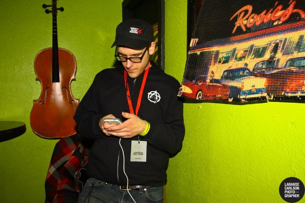 Eine Person mit einer Mütze und einem ID-Karte hält ein Mobiltelefon in der Hand und sieht auf dessen Bildschirm, mit einer Gitarre links und einem Plakat rechts.