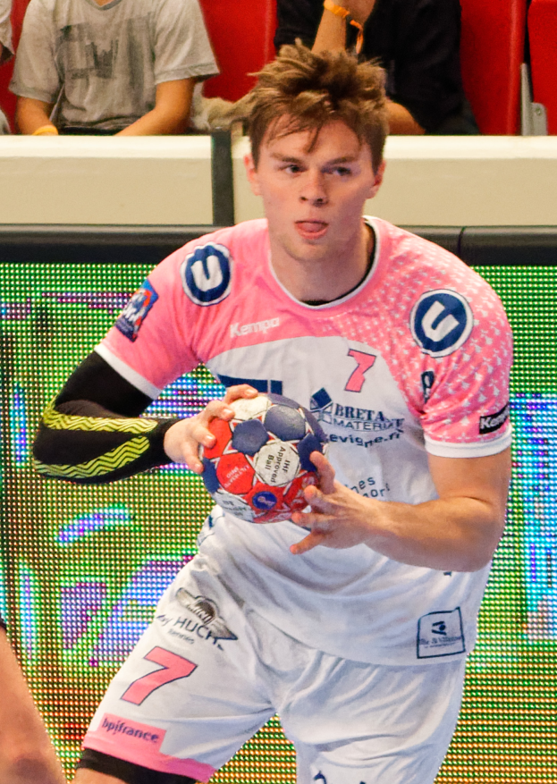 Ein Mann in einem rosa Hemd hält einen Fußball, mit der Hand eines anderen Menschen auf der linken Seite und ein paar Menschen im Hintergrund auf Stühlen sitzend.