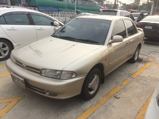 Ein goldfarbener Wagen auf einem Parkplatz neben anderen Autos, Gebäuden, Strommasten, Bäumen und einem klaren blauen Himmel.