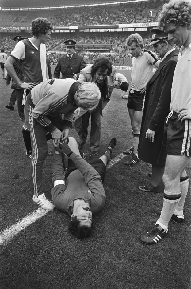 Schwarzes und weißes Bild eines Fußballspielers, der auf dem Boden liegt, während ihn eine Gruppe von Männern, darunter ein Schiedsrichter, in einem Stadion mit Zuschauern betreut.