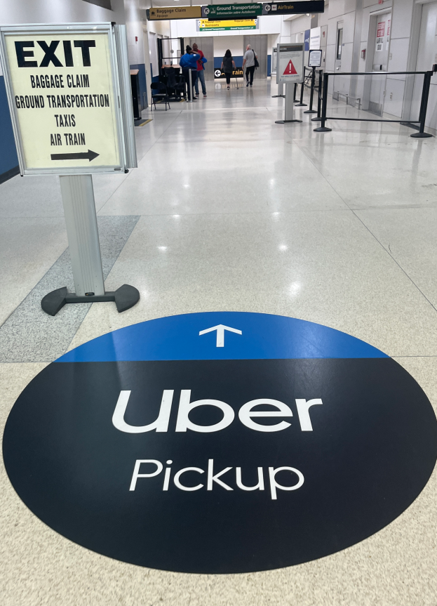 Ein Flughafen-Innere mit einem "Uber Abholen"-Schild auf dem Boden, Menschen im Hintergrund und verschiedene Gegenstände wie Bretter und Lampen.