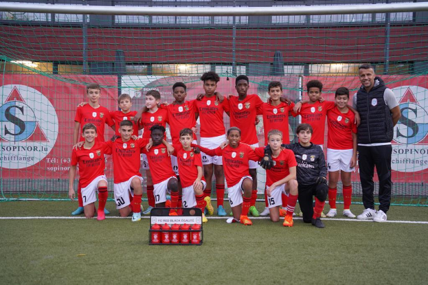 Eine Gruppe junger Männer in roten T-Shirts und schwarzen Shorts steht auf einem Fußballfeld neben einer Kiste roter Gegenstände, im Hintergrund Schilder mit der Aufschrift "FC Rot-Schwarz Eglaute - Champions League"