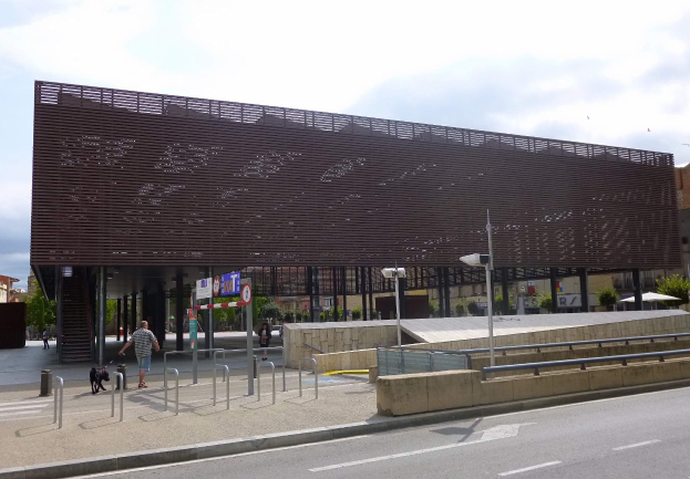Großes modernes Gebäude mit zahlreichen Fenstern, das als neuer Eingang zur Stadtzentrum dient, umgeben von Straßeninfrastruktur, Fahrzeugen, Fußgängern und Bäumen unter einem bewölkten Himmel.