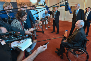 Ein Mann im Rollstuhl in Anzug und Krawatte von Reportern mit Kameras und Mikrofonen umgeben