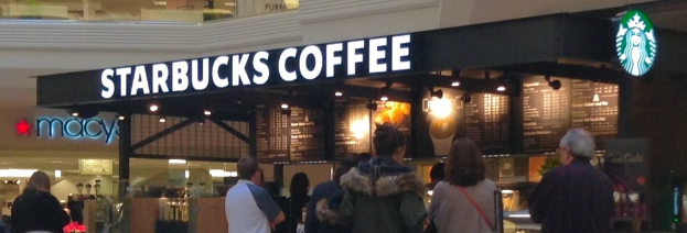 Menschen vor einem Coffeeshop mit Namen, Logo, Speisekarte und Lampen, mit Metallstangen und einem weiteren Geschäft daneben und einem Gang über dem Coffeeshop.