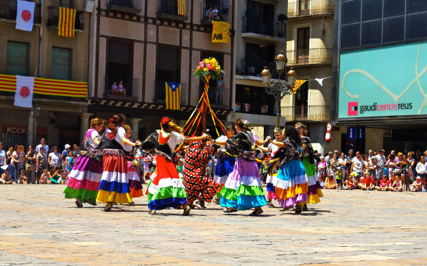 Eine Gruppe von Frauen in bunten Kleidern tanzt auf der Straße, umgeben von einer Menschenmenge, mit Gebäuden, Geländern, Balkonen, Fahnen, einem Laternenpfahl und einer Tafel mit Schrift im Hintergrund.