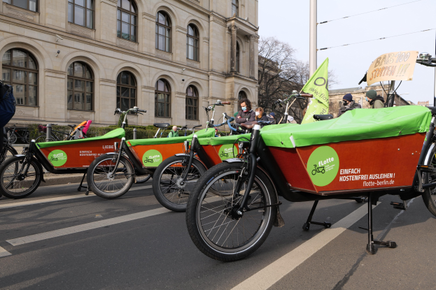 Gruppe von Fahrrädern, die an einer Straße geparkt sind, mit einer Person in der Nähe, Gebäuden und Bäumen im Hintergrund unter einem klaren blauen Himmel, mit einem Fahrrad-Sharing-Banner im Vordergrund.
