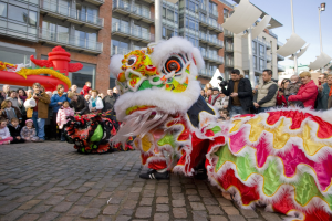 Ein lebendiges chinesisches Neujahrsfest in Amsterdam mit einer Löwen-Tanz-Show vor einer Zuschauermenge, einige halten Kameras, vor einem Hintergrund aus Gebäuden, Laternenmasten und einem klaren blauen Himmel.